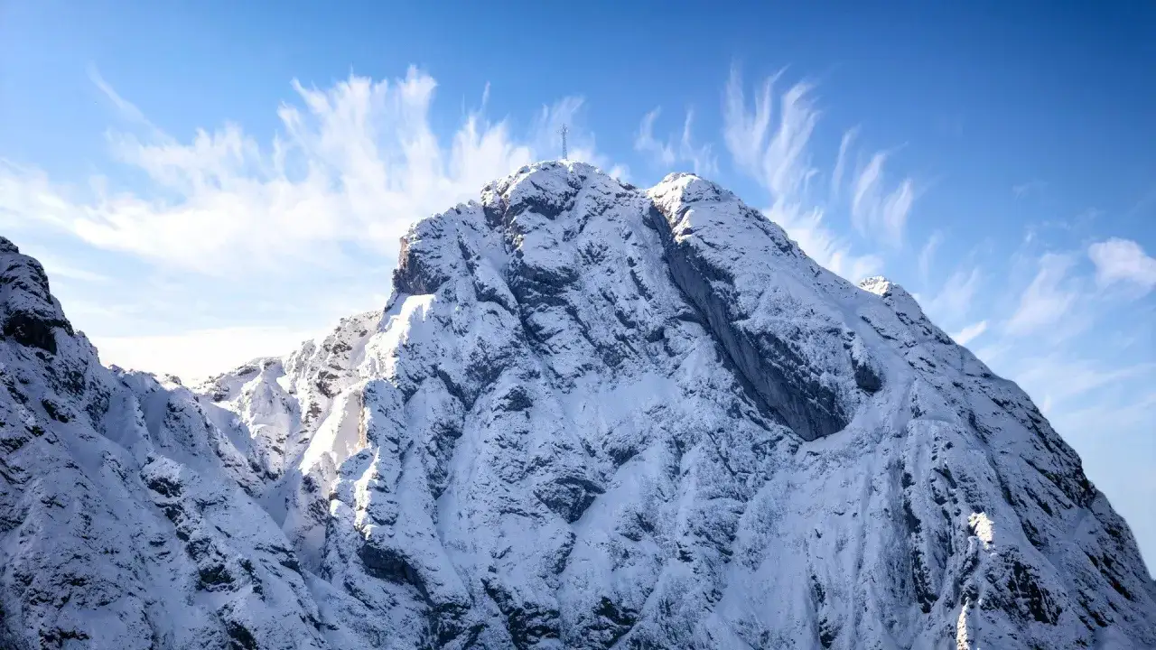 Pokryty śniegiem Giewont z krzyżem na szczycie, pod błękitnym niebem z pierzastymi chmurami. Sprawdź **Giewont mountain forecast** przed wyprawą.