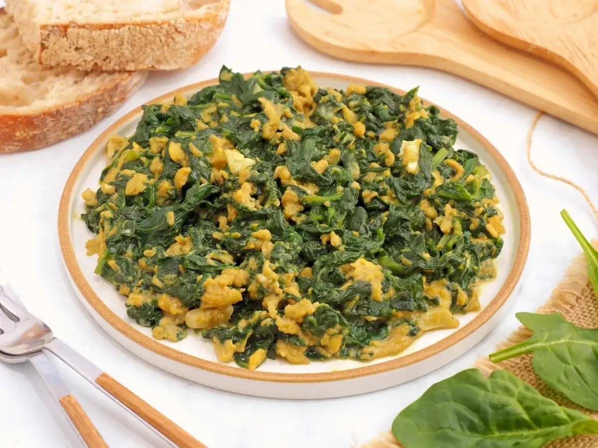 Delicioso revuelto de espinacas con huevo, servido en un plato blanco con borde marrón. Acompañado de rebanadas de pan rústico.