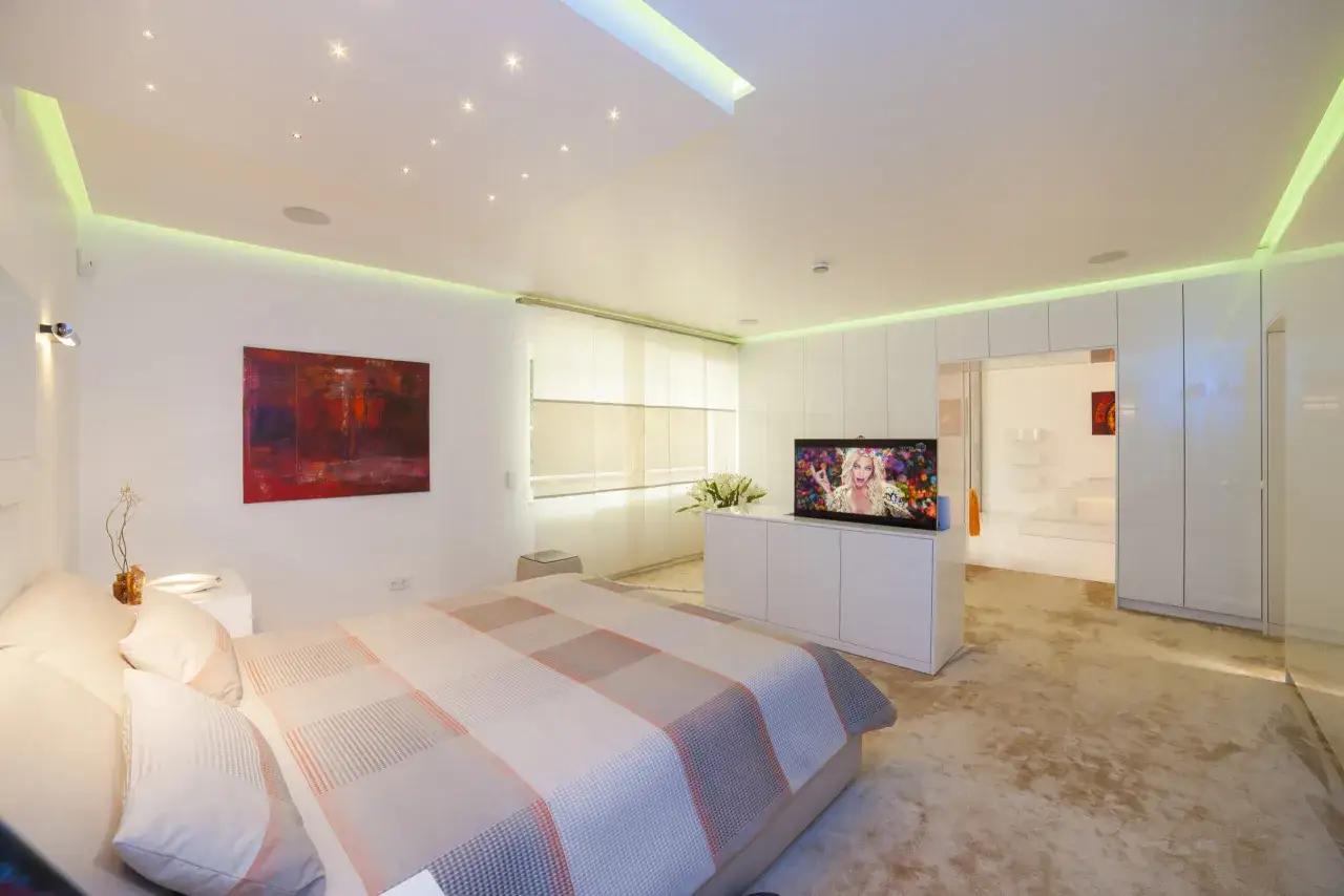 Modernes Schlafzimmer mit einem großen Fernseher im Schrank, der eine bunte Frau zeigt. Gemütliches Bett und dezente Beleuchtung.