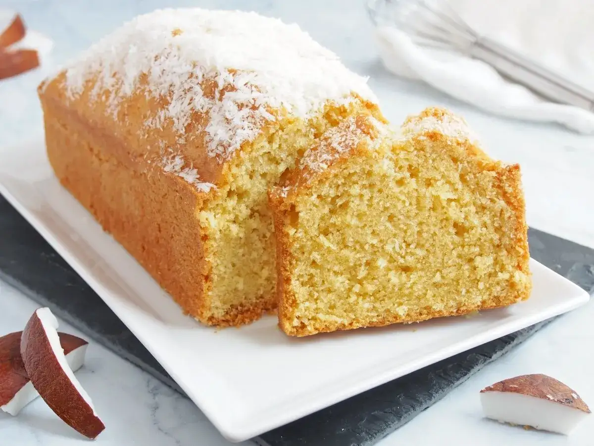 Bizcocho de coco esponjoso, cubierto con coco rallado, servido en plato blanco con trozos de coco fresco alrededor.