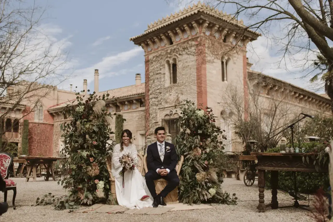 Novios sentados en sillas de mimbre, rodeados de exuberante vegetaci&oacute;n y flores, con una arquitectura antigua de fondo. Una hermosa decoracion boda boho.