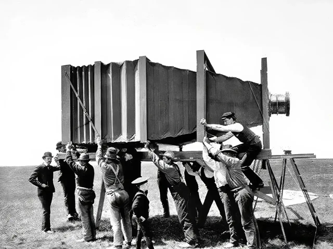 Grupa mężczyzn ustawia gigantyczny aparat, jeden z pierwszych aparatów fotograficznych. Widać jego harmonijkowy mieszek i obiektyw.