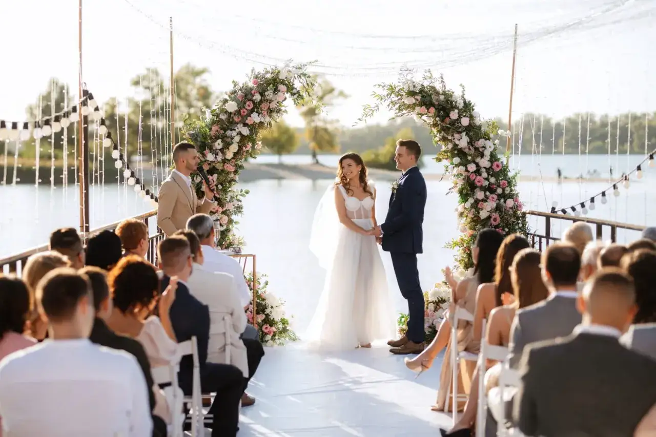 Un couple se tient la main sous une arche fleurie lors d'une **c&eacute;r&eacute;monie la&iuml;que ext&eacute;rieur**. L'officiant parle &agrave; des invit&eacute;s assis.
