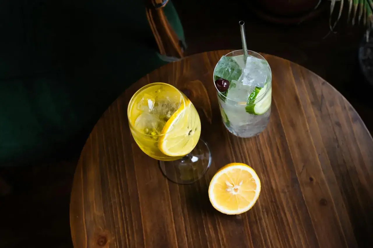 Dwa drinki na drewnianym stole. Jeden żółty z cytryną, drugi z ogórkiem i wiśnią. Skutki użycia taniego toniku do rzemieślniczego ginu: strata aromatu.