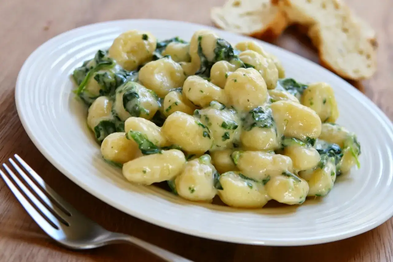 Pyszne makaron gnocchi w kremowym sosie ze szpinakiem, podane na białym talerzu. Obok widoczne kromki chleba.