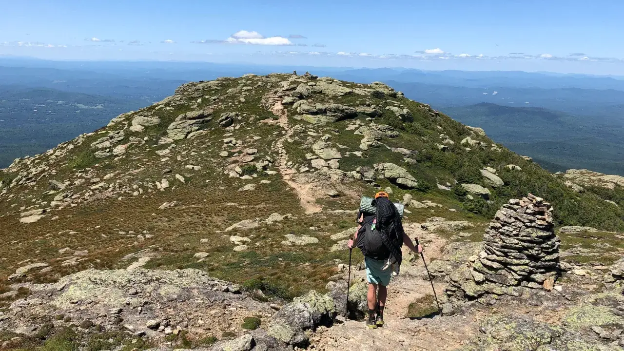 A hiker with a large backpack and trekking poles ascends a rocky, grassy mountain trail. The question of how long to hike 16 miles is on my mind as I take in the vast, blue mountain landscape.