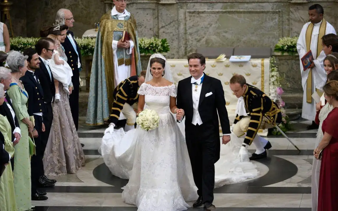 Madeleine och Chris går hand i hand under sitt bröllop. Bruden bär en spetsklänning och brudbukett, brudgummen frack.