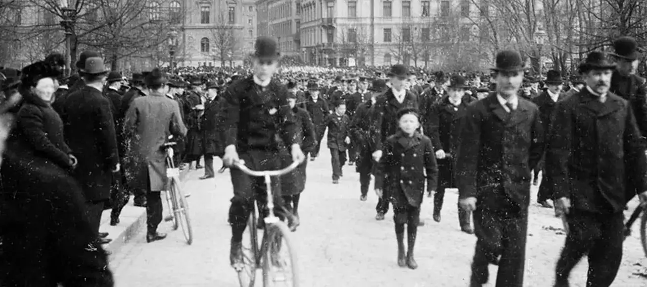 Folksamling i Stockholm 1900. Män i hattar och kostymer, en cyklist och en pojke i förgrunden.