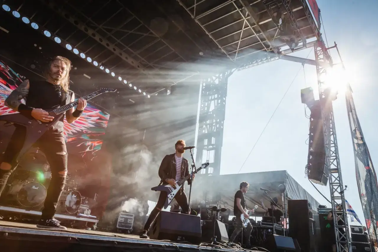 Drei Gitarristen einer Metal-Band spielen auf einer Bühne unter strahlendem Sonnenschein. Rauch und Lichteffekte erzeugen eine dramatische Atmosphäre.