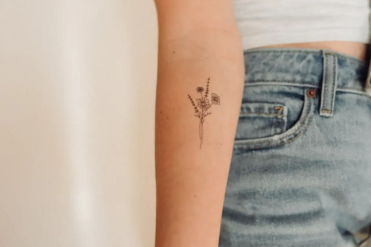 Ein zartes Blumenbouquet als minimalistische Tattoos auf einem Unterarm. Kleine Gänseblümchen und Lavendelzweige sind fein gezeichnet.