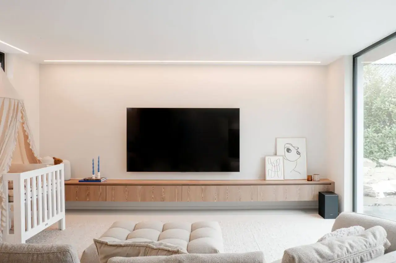 Un meuble TV suspendu, épuré et en bois, sous une grande télévision. Un lit de bébé est à gauche.