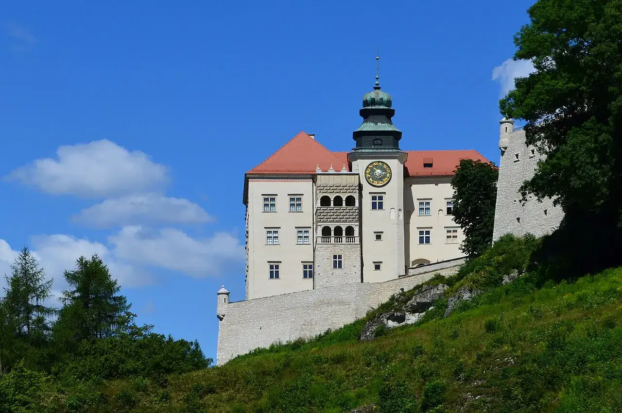 Zamek na pieskowej skale, z wieżą zegarową i czerwonym dachem, góruje nad zielonym zboczem.