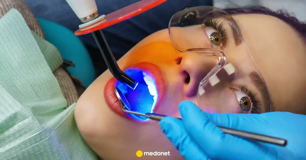 Zabieg lakowania zębów uśmiechniętej pacjentki. Dentysta utwardza światłem lampy specjalny materiał.
