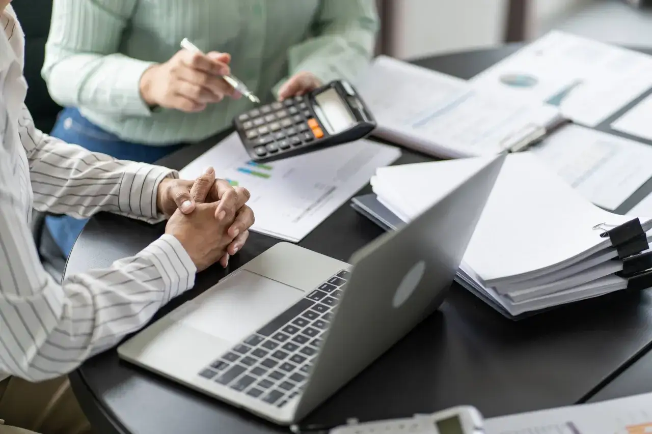Two people review compiled financial statements, using a calculator and laptop to analyze data.