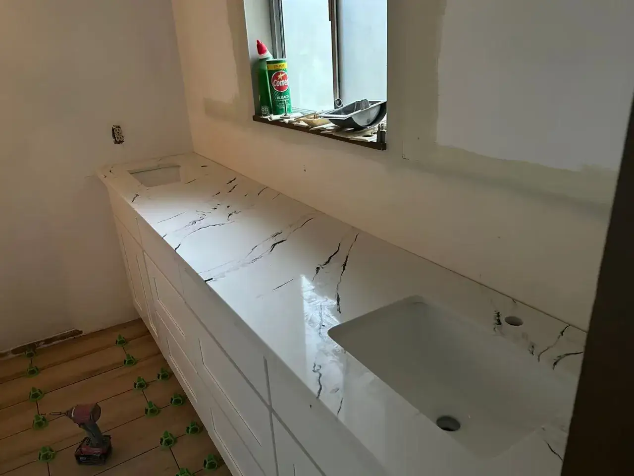 Łazienka w trakcie remontu. Biały blat z czarnymi żyłkami, imitujący konglomerat carrara, z dwoma umywalkami.