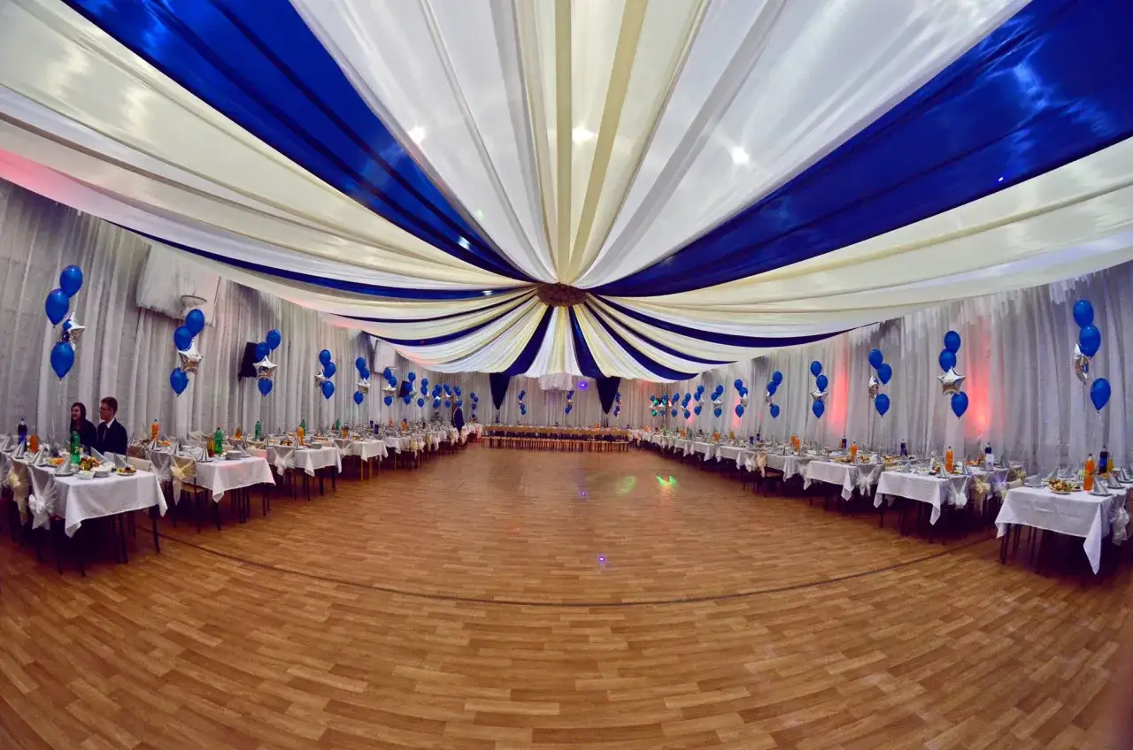 Sala bankietowa udekorowana białymi i niebieskimi tkaninami, balony, stoły nakryte na komers.