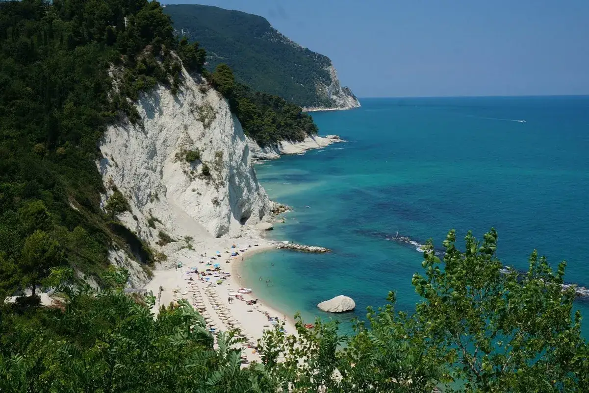 Spiaggia dei frati con scogliere bianche, mare turchese e alberi verdi. Ombrelloni e persone godono del sole.