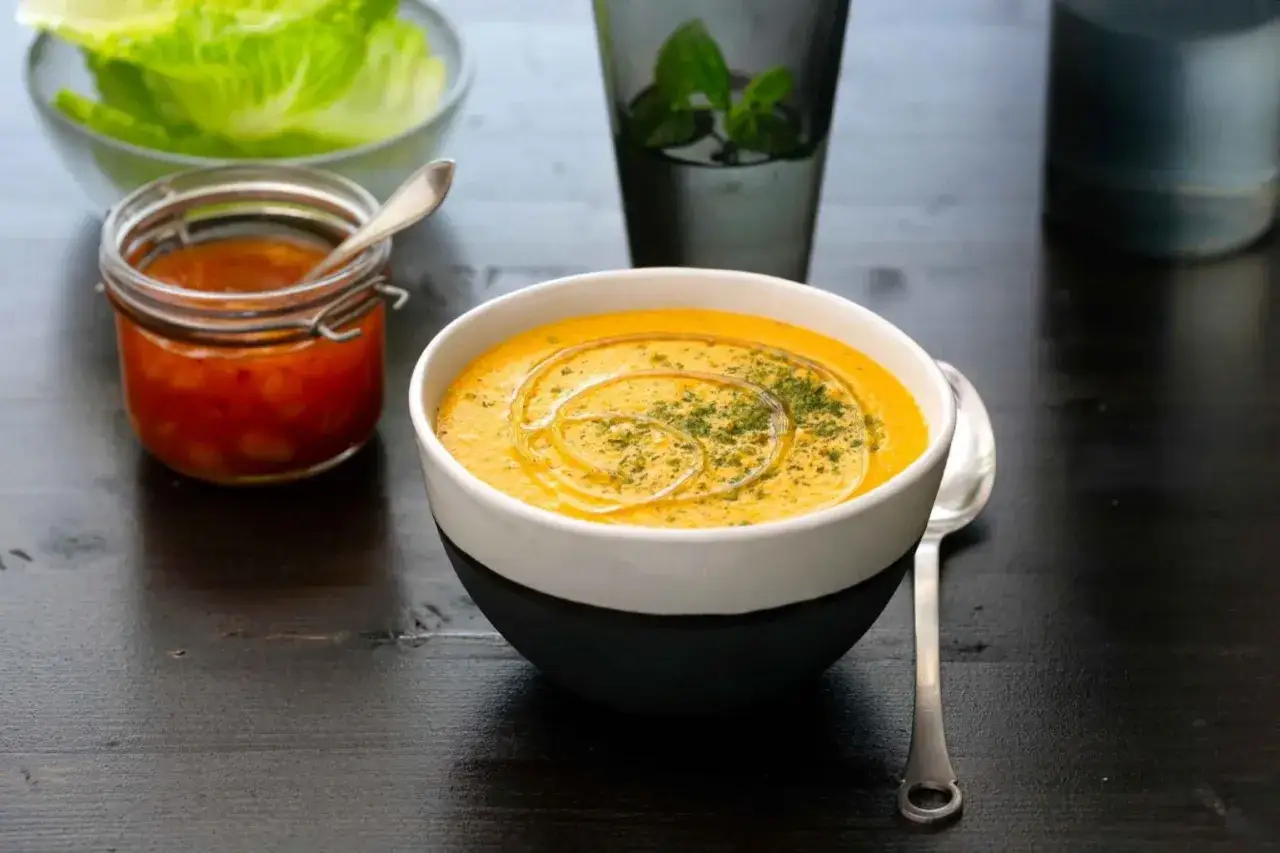 En kr&auml;mig linssoppa med en virvel av olja och &ouml;rter. Bredvid st&aring;r en burk med en r&ouml;d r&ouml;ra och en sk&aring;l med salladsblad. Ett enkelt linssoppa recept.