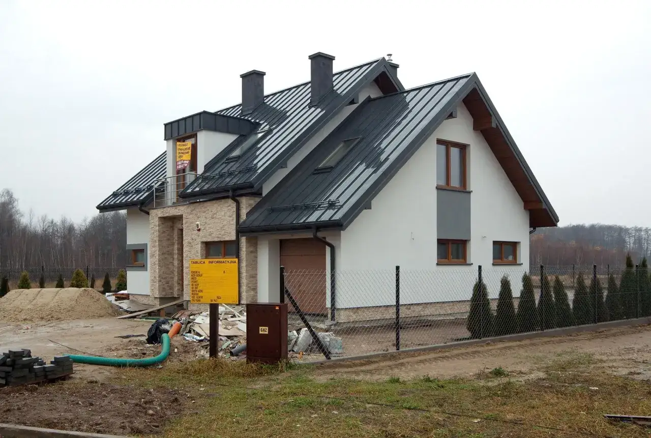 Nowy dom w budowie, trwa wykonanie przyłącza elektrycznego. Widać koparkę, materiały budowlane i tablicę informacyjną.