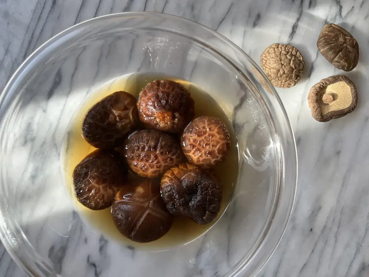 Get ready for the amazing shiitake wirkung! Several rehydrated shiitake mushrooms swim in a clear broth in a glass bowl, with dried shiitakes nearby.