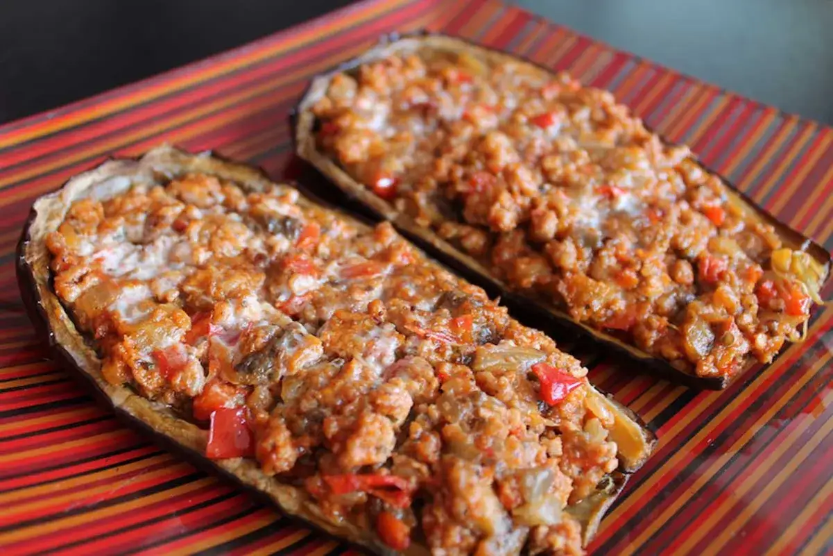 Berenjenas rellenas de carne picada, con trozos de pimiento rojo y queso gratinado, servidas en un plato a rayas.
