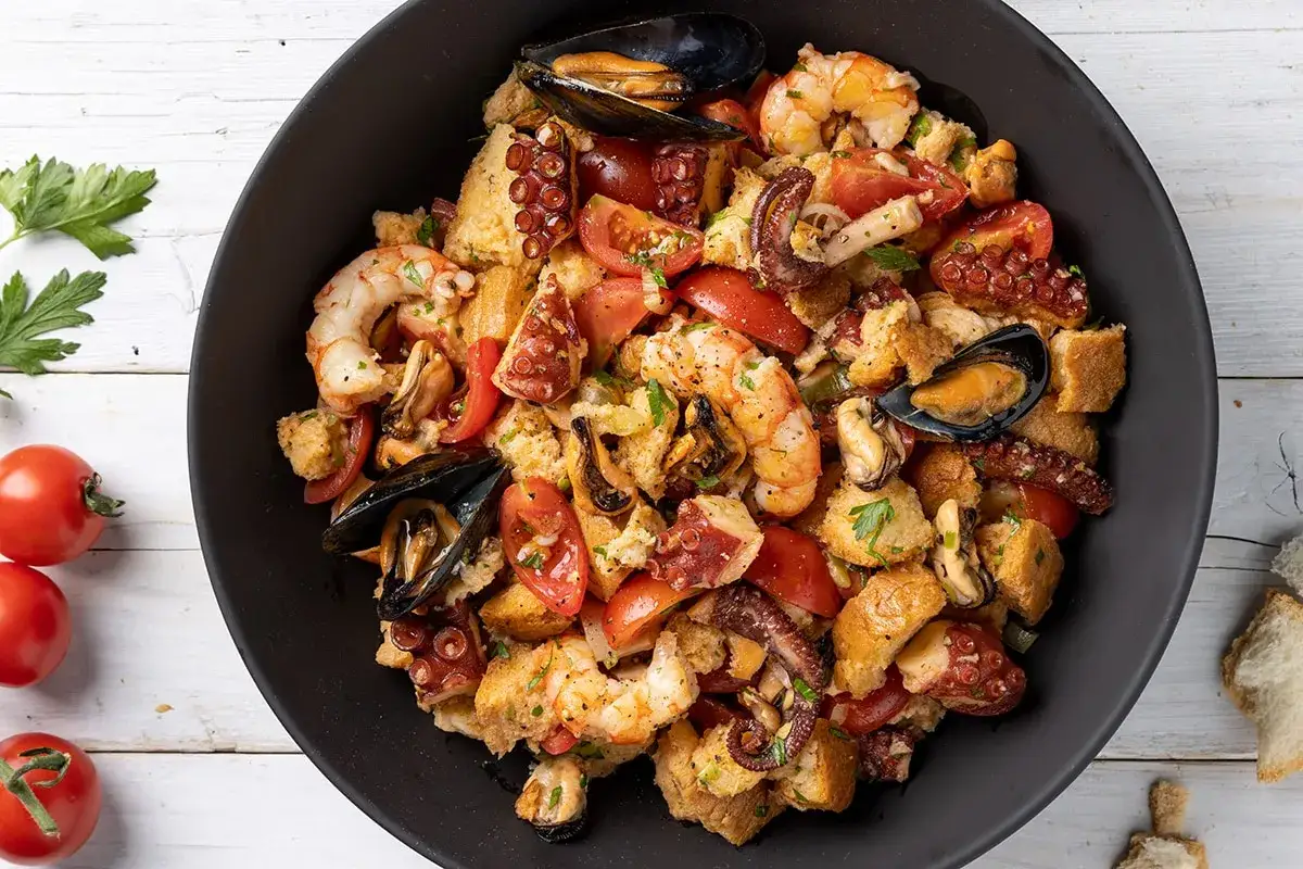 Panzanella di mare ricca di gamberi, polpo, cozze e pomodori, con crostini di pane croccante. Un piatto estivo che sa di mare.