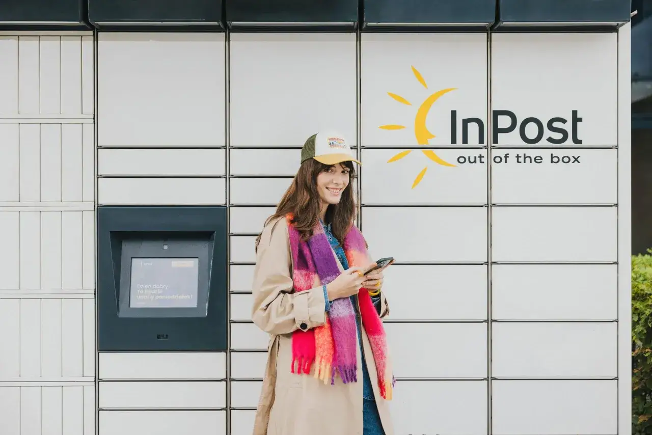 Kobieta z telefonem przy paczkomacie InPost. Zastanawiasz się, ile prądu zużywa paczkomat? To urządzenie jest energooszczędne.