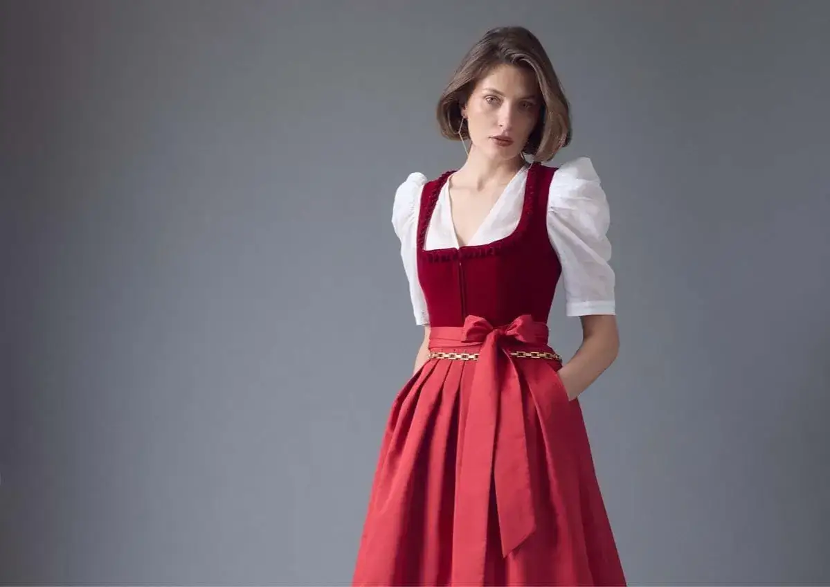 Frau im roten Dirndl mit wei&szlig;er Bluse. Die Dirndlsch&uuml;rze aus Seide ist mit einer goldenen Kette und einer Schleife verziert.