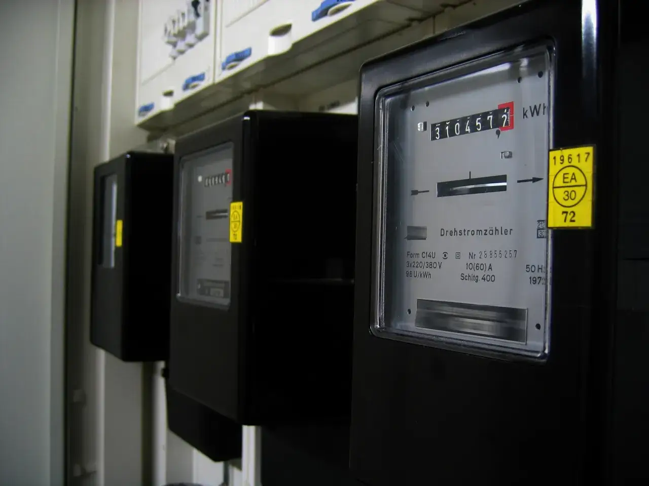 Szereg czarnych liczników energii elektrycznej, każdy z wyświetlaczem pokazującym zużycie. Na jednym z nich widoczny jest żółty znaczek z napisem