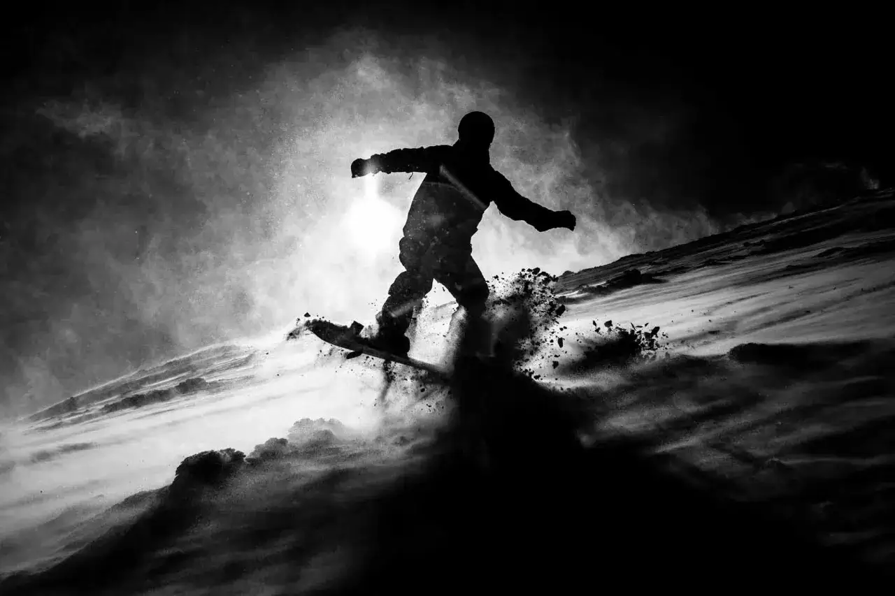 Silueta snowboardzisty w locie, z chmurami śniegu wokół. Dramatyczne **światło i cień w fotografii** podkreślają dynamikę ruchu.