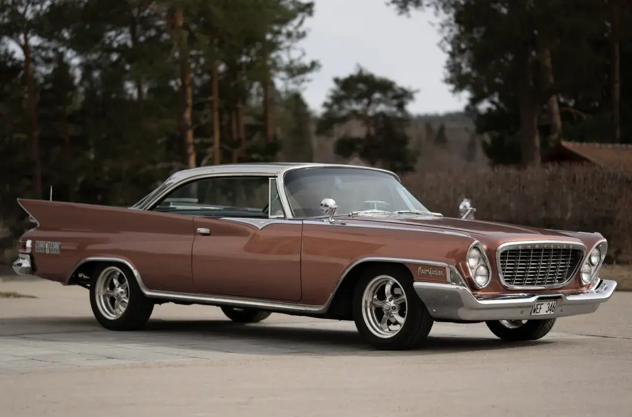 Brun Chrysler New Yorker med kromdetaljer och fenor, parkerad utomhus bland träd.