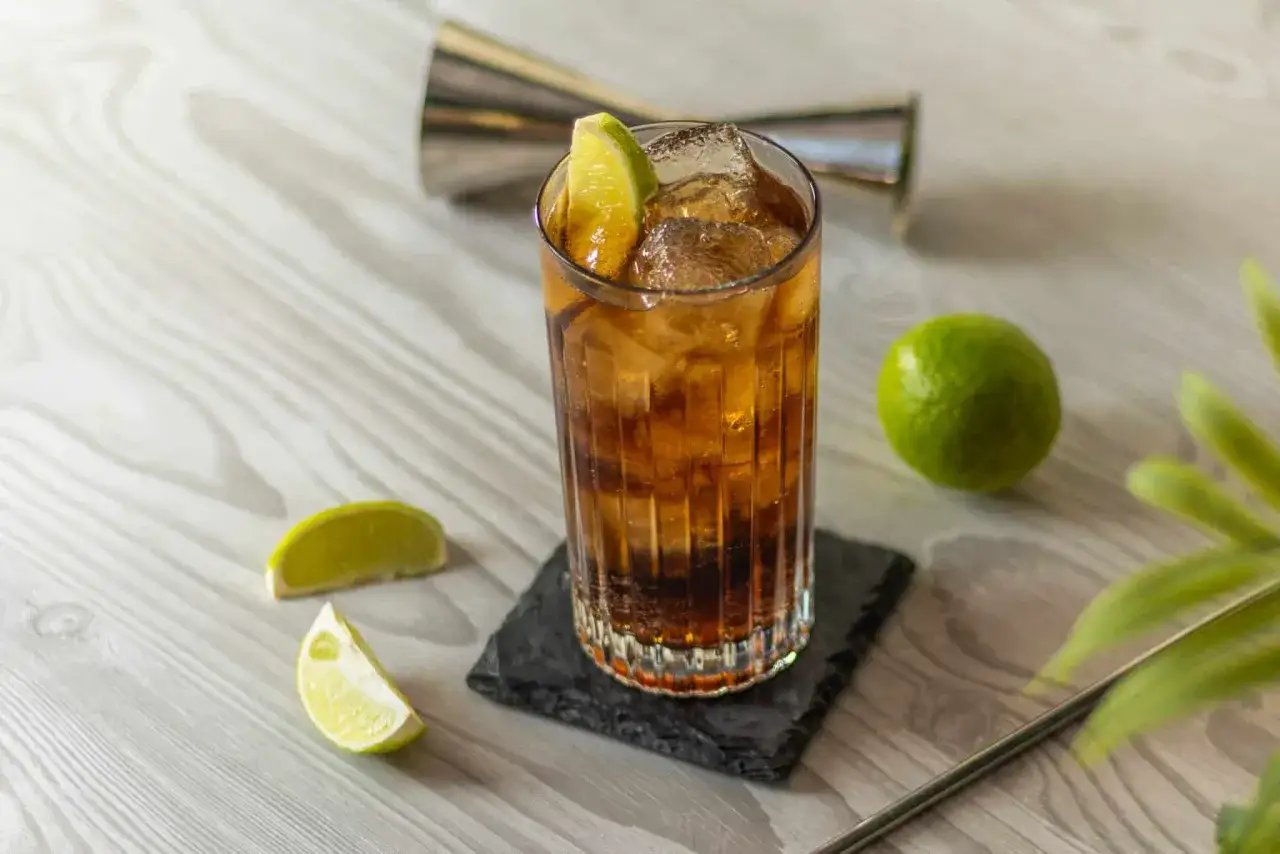 Orzeźwiające whisky z colą, ozdobione plasterkami limonki, z kostkami lodu i metalową shakerką w tle.
