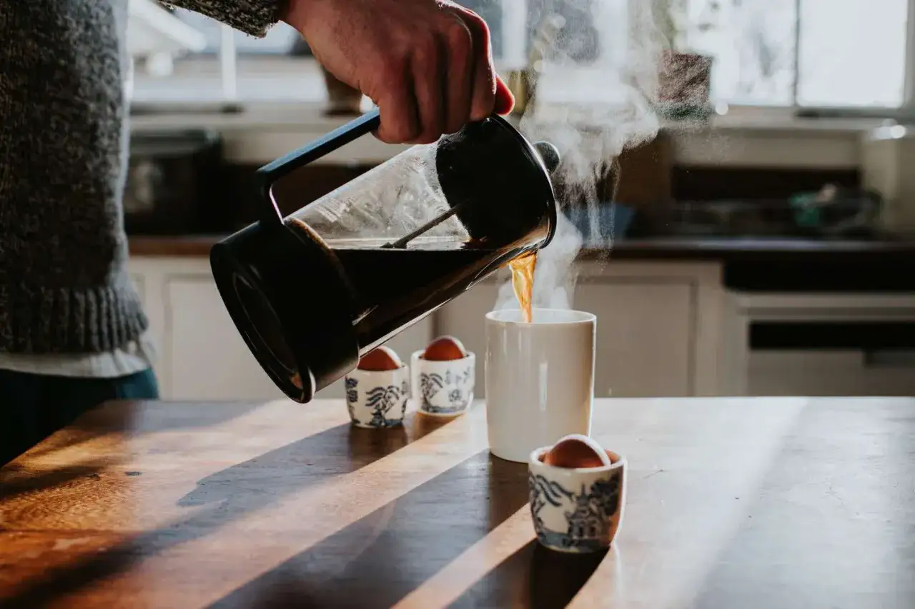 Eine Hand gie&szlig;t dampfenden Kaffee aus einer French Press in eine Tasse. Daneben stehen Eierbecher mit Eiern. Manche sagen, Kaffee ohne Filter sei ungesund.