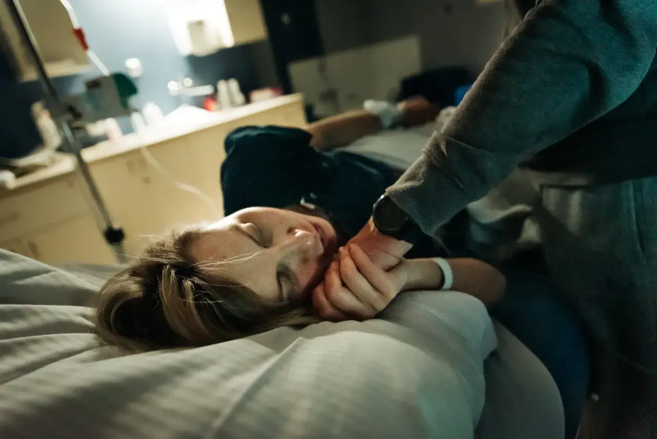 Kobieta odpoczywa w łóżku szpitalnym, trzymając dłoń bliskiej osoby. W tle widać sprzęt medyczny, sugerujący pobyt na **OIOM**.