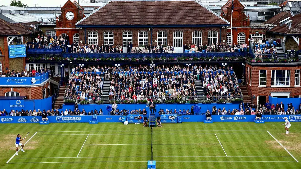 Jugadores en el torneo de Queen's Club, con gradas llenas de espectadores y el ic&oacute;nico edificio de ladrillo rojo de fondo.