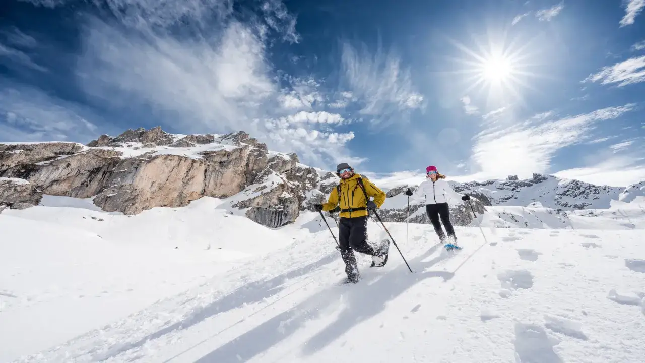 Coś takiego jak ciaspole cosa sono? Due persone camminano sulla neve con le ciaspole, godendosi un paesaggio montano soleggiato.