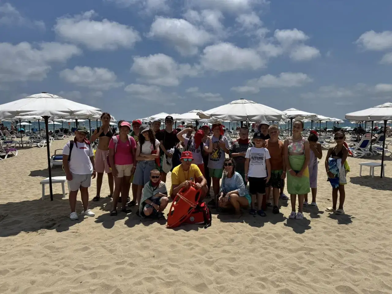 Grupa dzieci i opiekunów na plaży w Bułgarii. Widać parasole, leżaki i morze. Takie są wakacje na obozach w Bułgarii.