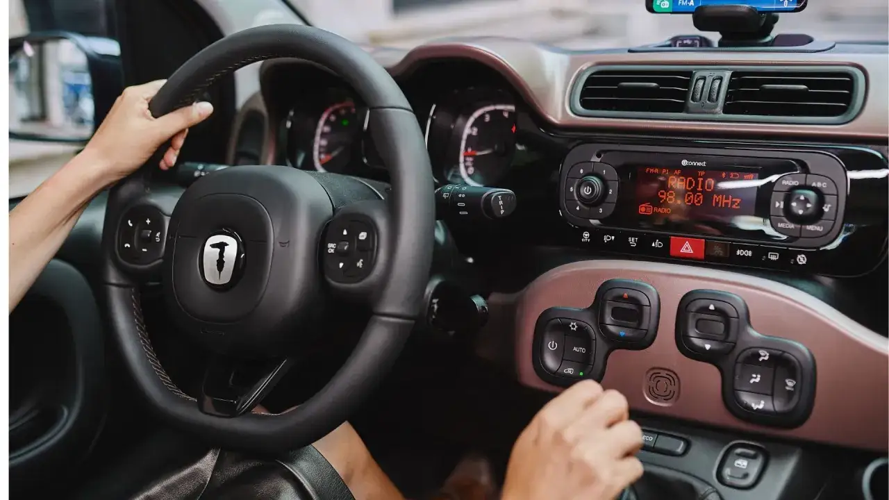 Mani sul volante di una Fiat Panda a gas, con il cruscotto e la radio accesi.