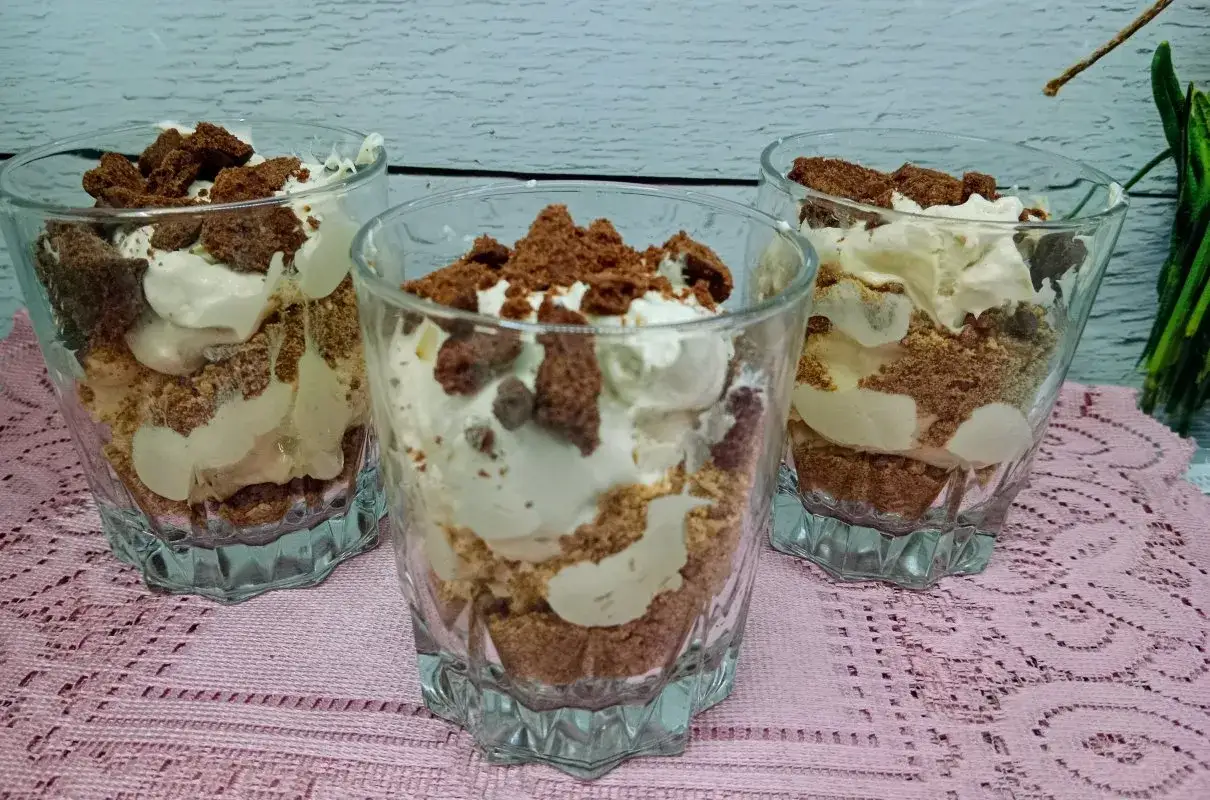 Trzy pucharki z biszkoptem z serkiem mascarpone i bitą śmietaną, posypane okruszkami ciasta.