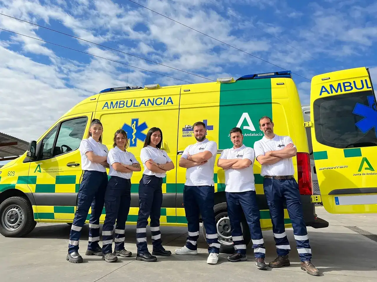 Equipo de fp t&eacute;cnico emergencias sanitarias posa orgulloso junto a una ambulancia amarilla y verde bajo un cielo azul con nubes.