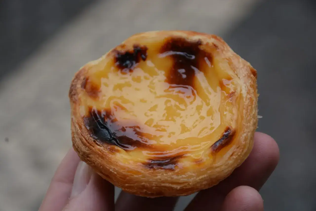 En hand h&aring;ller en gyllene, karamelliserad pastel de nata med kr&auml;mig fyllning.