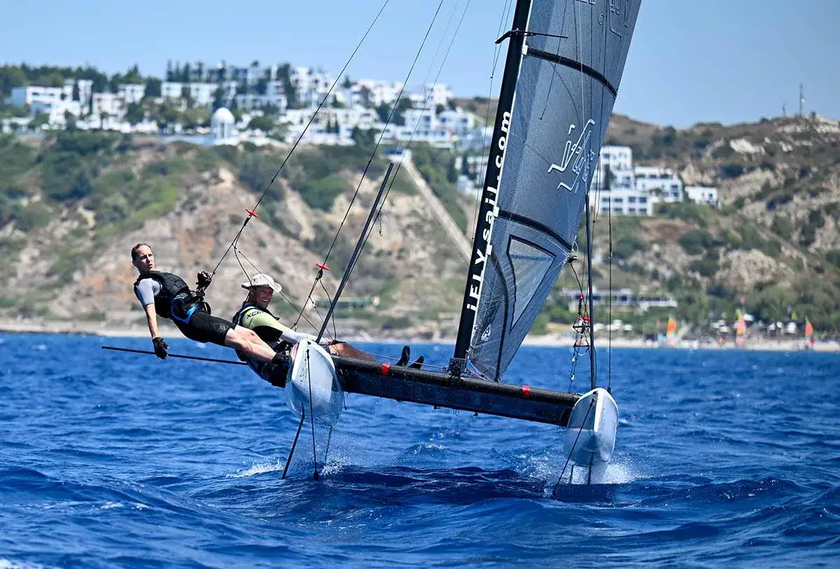 Zwei Männer auf einem Katamaran, der mit Foil surfen ohne segel über das Meer gleitet. Im Hintergrund eine Küste mit weißen Häusern.