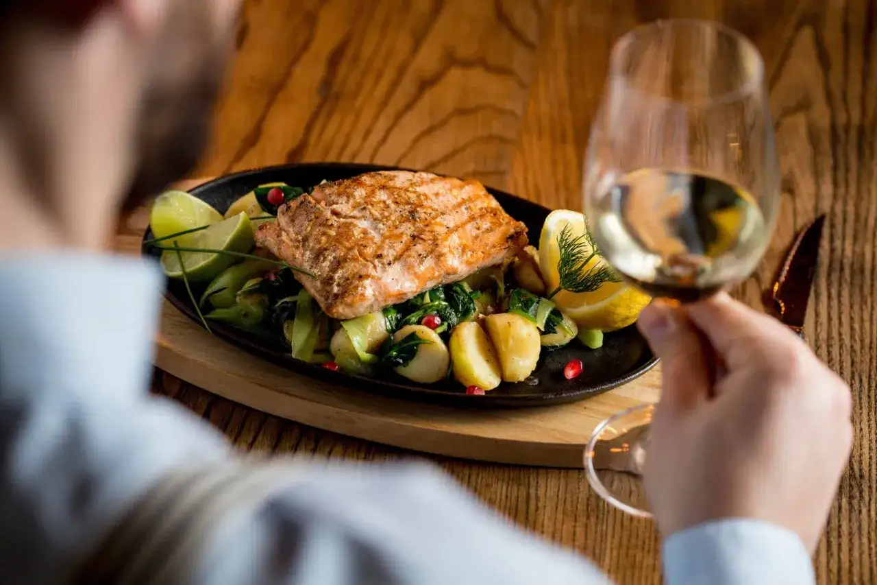 En man njuter av en utsökt laxrätt med potatis, grönsaker och citron, perfekt ackompanjerad av ett glas vitt vin till Sandefjordsås.