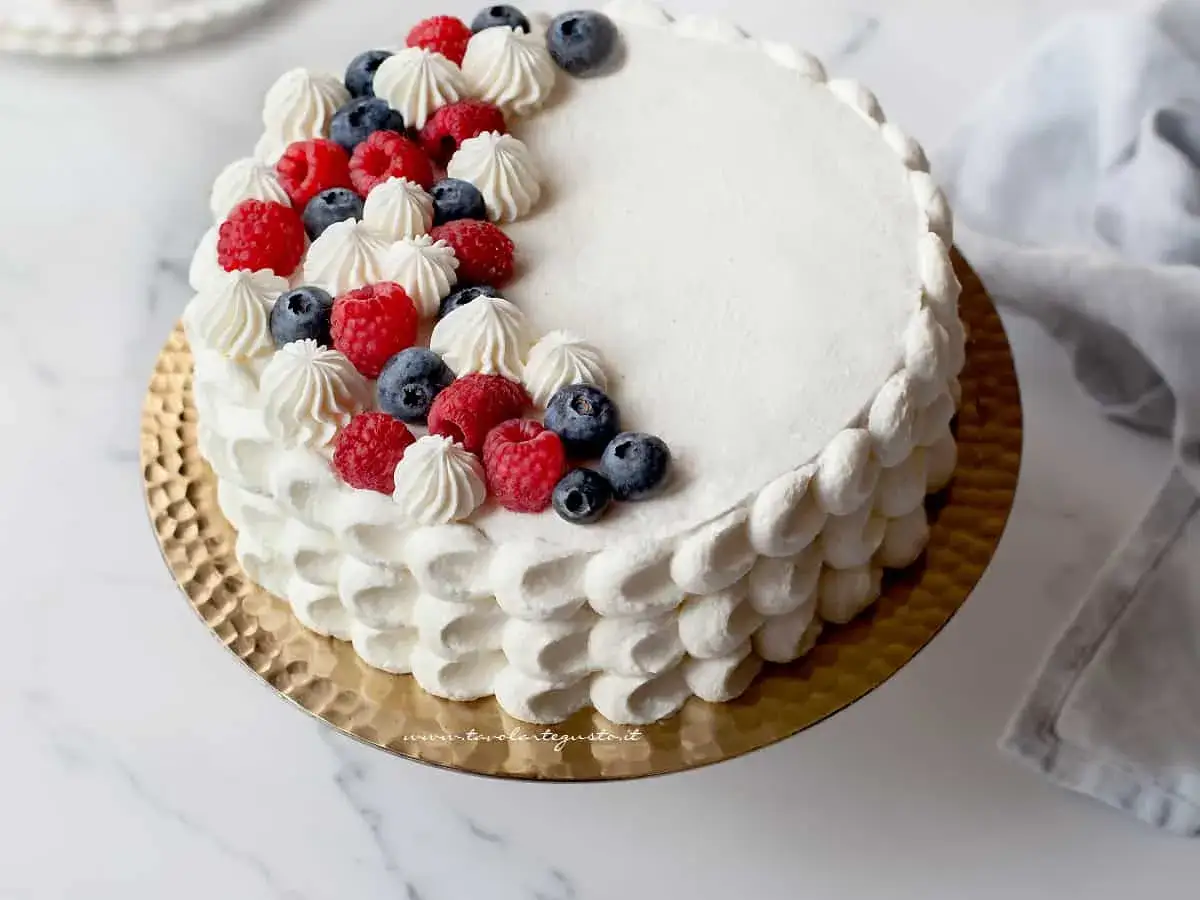 Torta compleanno pan di spagna con panna, lamponi e mirtilli, decorata con ciuffi di panna e frutta fresca.