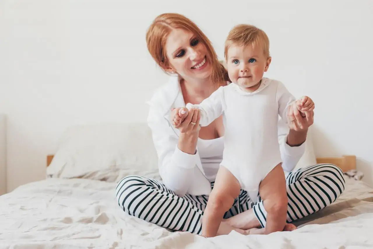 Uśmiechnięta mama pomaga 8-miesięcznemu dziecku w pierwszych krokach. Niepokojące sygnały rozwoju niemowlaka mogą być subtelne.