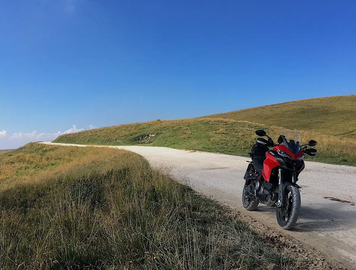Ducati rossa su strada sterrata, con un paesaggio collinare e cielo azzurro. Un invito a esplorare il passo fittanze.