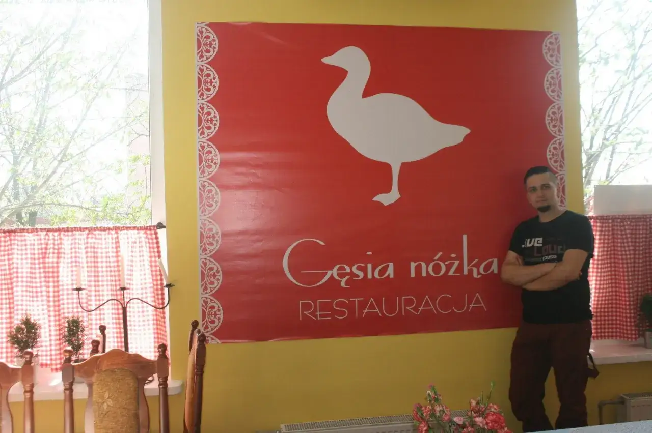Mężczyzna stoi obok wielkiego czerwonego banera z białym gęsią i napisem 