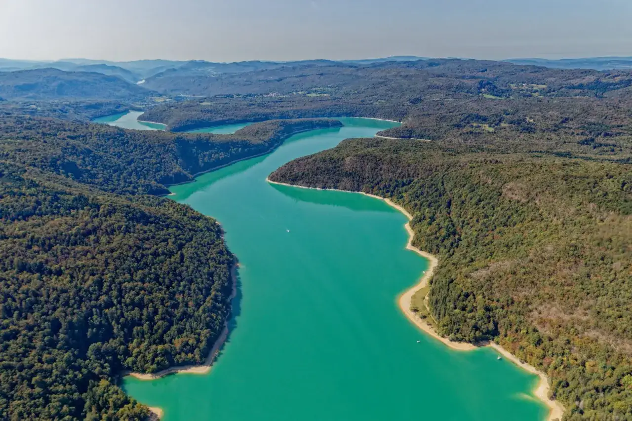 Vue aérienne d'un lac aux eaux turquoise, serpentant entre des collines boisées du lac Jura. Un petit bateau navigue paisiblement.