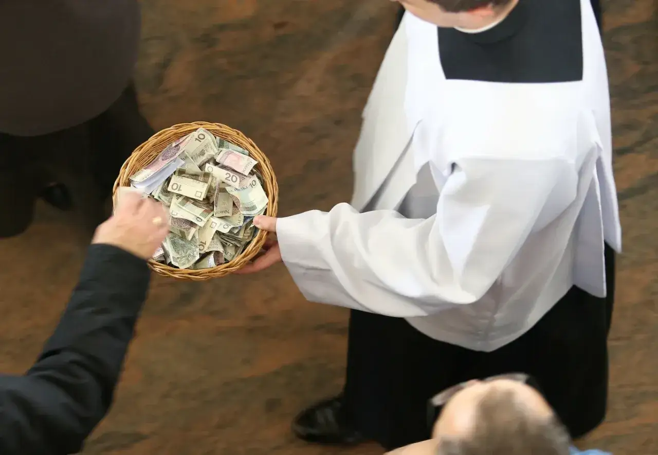 Ksiądz zbiera datki do koszyka. Widać polskie banknoty, które są częścią podatek na kościół.