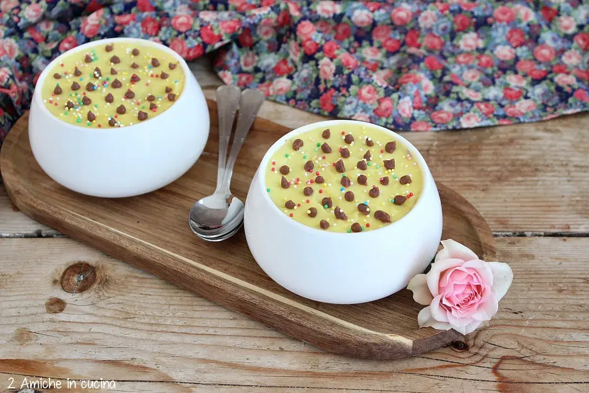 Deliziosa crema pasticcera senza uova, guarnita con gocce di cioccolato e zuccherini colorati, servita in ciotole bianche su un tagliere di legno.